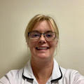 Headshot photo of Kathy Cann smiling at the camera. She has blonde hair tied back in a pony tail, is wearing glasses and a white clinical uniform